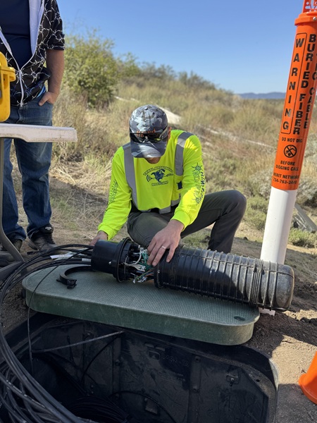 TBB instructor kneels next to an open fiber vault in the ground examining and exposed cylindrical splice case