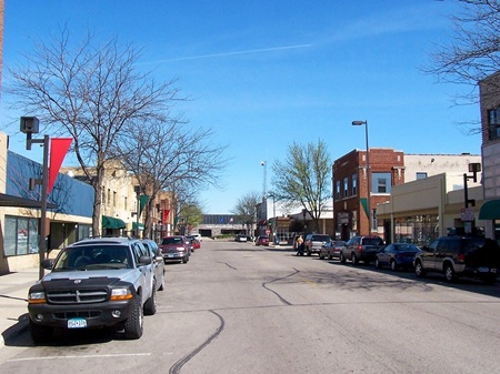 Daytime in Downtown Willmar shows a row of buildings on two sides of the street none of whom are more than 2 stories. Park car line each side of the tree-lined street