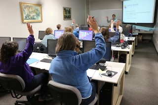 Seniors in computer class A classroom full of seniors raising their hands as the instructor stands at the front of the room