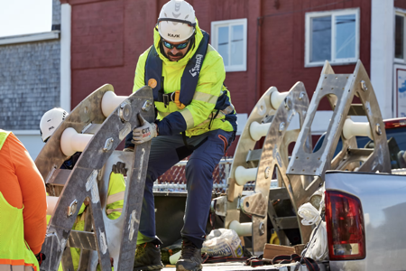 A worker wearing a hard hat and bright yellow shirt pulls equipment into back of pick up truck