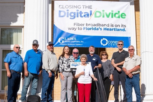 Nearly a dozen Glades Electric Cooperative and Conexon officials stand in front of a sign that says "Closing the Digital Divide"