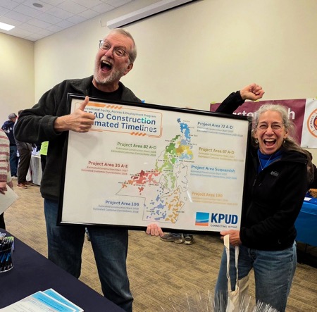 Two Kitsap PUD officials celebrate getting BEAD grant holding a sign announcing construction deadlines