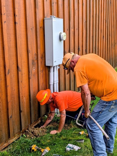 Two installation technicians crouch near a fence in a yard threading a fiber wire under the fence 
