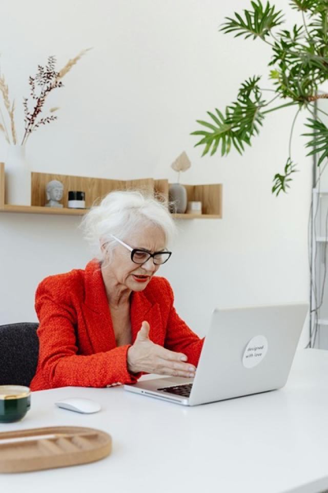 An older woman wearing a red suit jacket looks distressingly at her laptop, motioning with her hand toward the screen