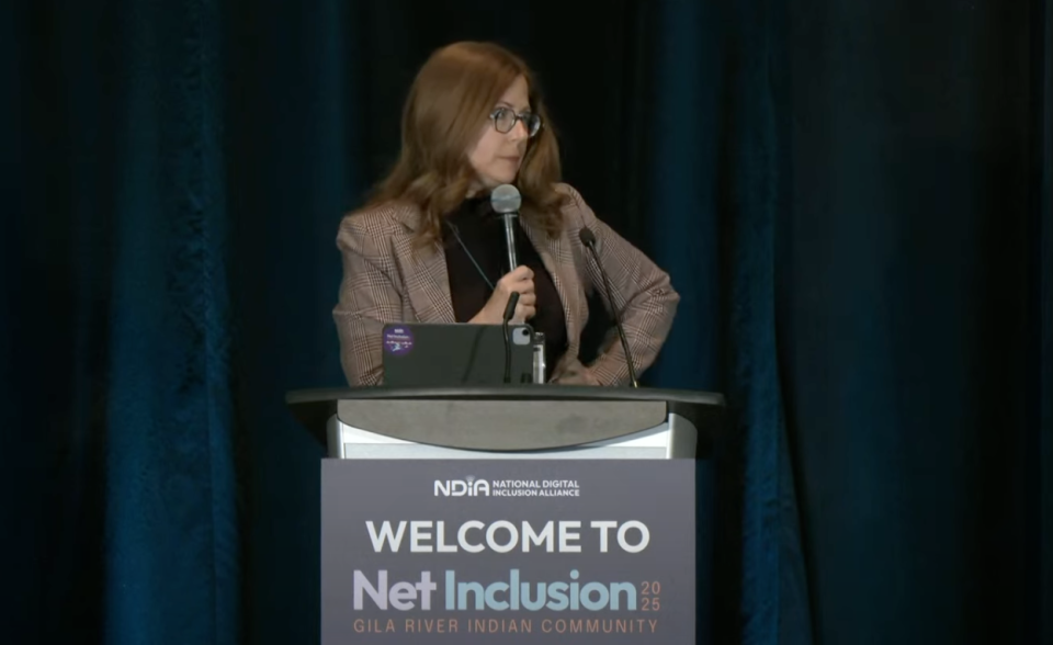 NDIA executive director Angela Siefer holds mic standing behind podium addressing Net Inclusion audience
