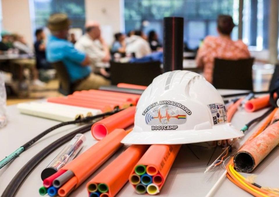 A hard says "Tribal Broadband Bootcamp" atop fiber conduit with a group of people seated in the blurred background