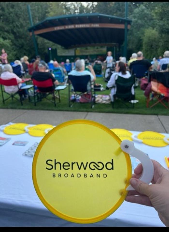 An employee holds up a Sherwood Broadband gift emblazoned with its name with dozens of residents sitting in on a park lawn listening to local concert