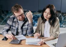 Couple frets over bills at a table. The man is holding a cup of coffee as he looks concerned about the bill his wife is showing him